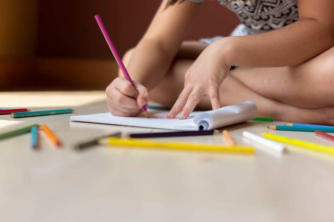 A child seated on the floor using colored pencils to draw on paper, showcasing creativity.